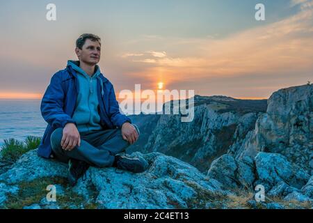 Tourist Mann in einer blauen Jacke sitzt auf einem Felsen hoch in den Bergen, über den Wolken und genießt den Sonnenuntergang Copy Space. Das Konzept der Ruhe, Stille Stockfoto