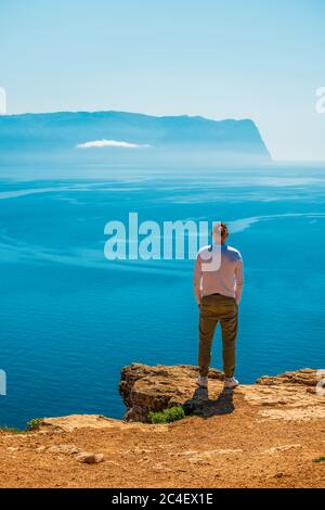 Junger stilvoller Mann junger Mann steht an der felsigen Küste mit Blick auf blaues azurblaues ruhiges Meerwasser an sonnigen Tagen. Sommerferienkonzept Cliff ragt aus Stockfoto