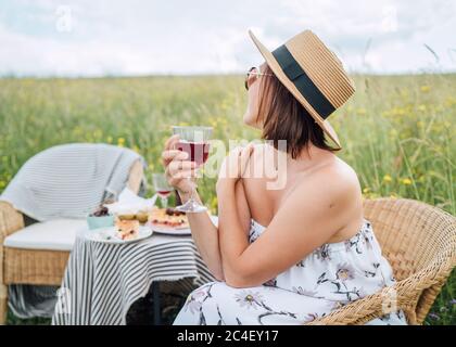 Junge Frau in Sonnenbrille und Strohhut gekleidet leichte Sommerkleid sitzen in Rattan Stuhl auf der hohen grünen Wiese neben Picknick-Tisch fröhlich, Stockfoto