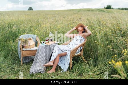 Junge Frau gekleidet leichte Sommerkleid und Strohhut sitzen in einem Rattan-Stuhl auf der hohen grünen Wiese und genießen den Moment in der Nähe des Picknicks Stockfoto