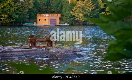 Retirement Living - zwei Muskoka Stühle sitzen auf einem felsigen Ufer mit Blick auf einen ruhigen See mit Bäumen und einem Boot Stockfoto