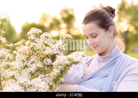 Mutterschaft, Pflege, Säuglinge, Sommer, Elternkonzept - Junge schöne Mutter mit einem Neugeborenen in einem Tragetuch geht und schaut auf ein Kind im Hintergrund Stockfoto