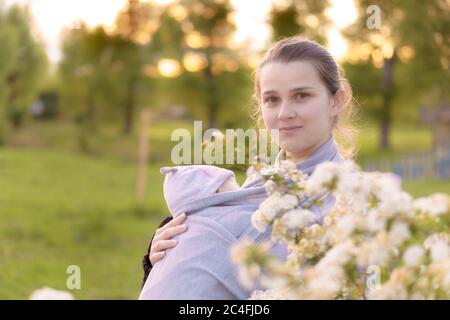 Mutterschaft, Pflege, Säuglinge, Sommer, Elternkonzept - Junge schöne Mutter mit einem Neugeborenen in einem Tragetuch geht und schaut auf ein Kind im Hintergrund Stockfoto