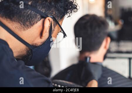 Mulatto Barbier mit einer Gesichtsmaske und Brille schneiden Haare auf Hals und Nacken des Mannes in Friseurladen mit professionellen Clipper-Tool. Elektrische Trimmer Maschine Cu Stockfoto