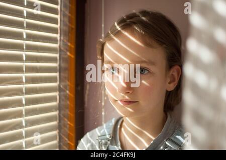 Das Portrait eines kleinen Mädchens mit langen blonden Haaren und blauen Augen sitzt am Fenster mit Jalousien während eines sonnigen Tages zu Hause Stockfoto