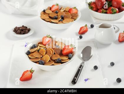 Süße hausgemachte Schale mit winzigen Pfannkuchen mit Schokolade, Honig, Erdbeeren und Heidelbeeren auf weißem Tisch. Nahaufnahme.Horizontal, selektiver Fokus. Stockfoto