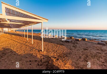 Menschenleerer Strand in der Nebensaison. Holzliegen am öffentlichen Sandstrand am Meer. Am Ufer und Steine im Wasser. Düsterer Himmel. Das Konzept von Stockfoto