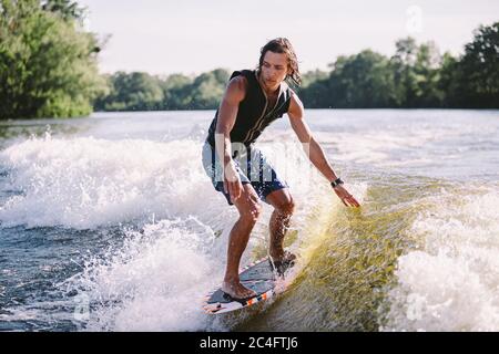 Schöne langhaarige blonde Wakesurf in Weste an Bord entlang Wellen des Sees. Sportliche männliche Sportler wacht im Sommer auf dem Fluss surfen. Wakesurf Wasser Stockfoto