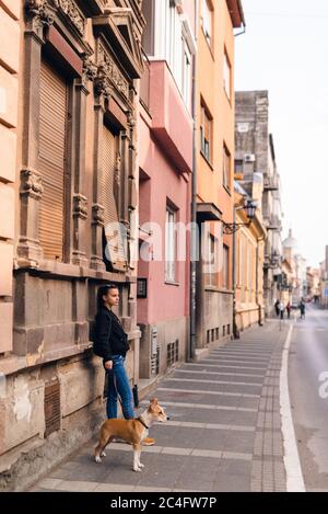 Ein schönes kaukasisches Mädchen mit Sonnenbrille steht neben ihrer Hunderasse American Staffordshire Terrier vor einem alten Haus auf der Straße Stockfoto