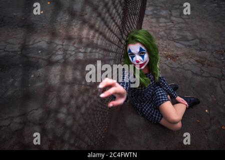 Greenhaired Mädchen in gechert Kleid mit Joker Make-up sitzt in der Nähe von Draht-Mesh-Zaun auf dem Boden. Weitwinkelaufnahme. Stockfoto