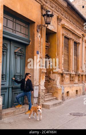 Ein schönes kaukasisches Mädchen mit Sonnenbrille steht neben ihrer Hunderasse American Staffordshire Terrier vor einem alten Haus auf der Straße Stockfoto