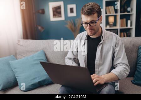 Glücklicher Kerl mit Laptop, während er auf der grauen Couch sitzt Stockfoto