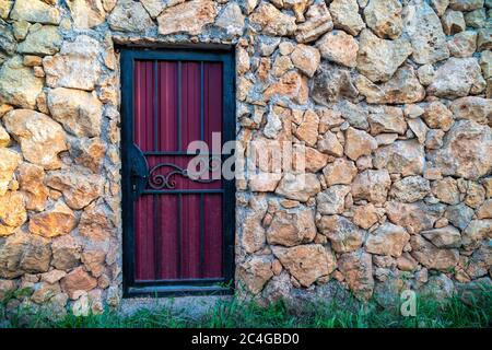 Eine violette Metalltür mit einem schwarzen Türknopf. Die Tür ist in einer Felswand. Hintergrund aus Kalkstein Mauerwerk. Die Mauer ist aus Wildstein. Der Stockfoto
