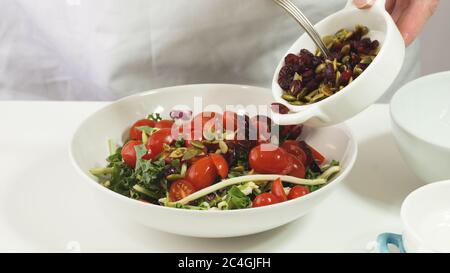 Zubereitung von Gemüsesalat. Frau legt Kürbiskerne und getrocknete Preiselbeeren in eine Schüssel mit verschiedenen Salatblättern und Tomaten, dicht am weißen Backg Stockfoto