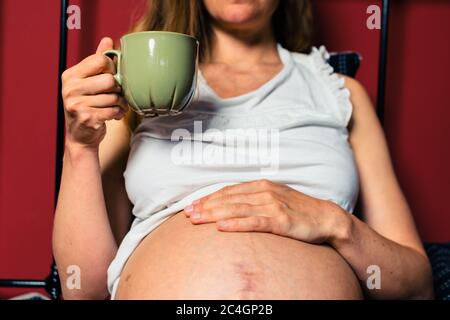 Eine junge schwangere Frau trinkt aus einem Becher im Bett Stockfoto