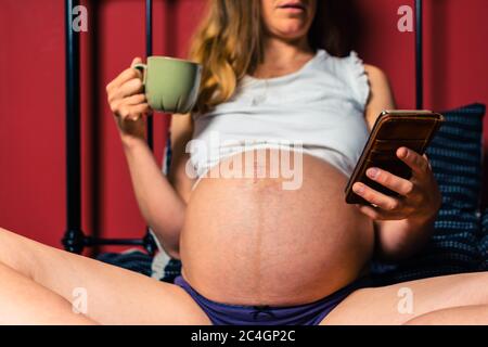 Eine junge schwangere Frau trinkt aus einem Becher und benutzt ihr Smartphone im Bett Stockfoto