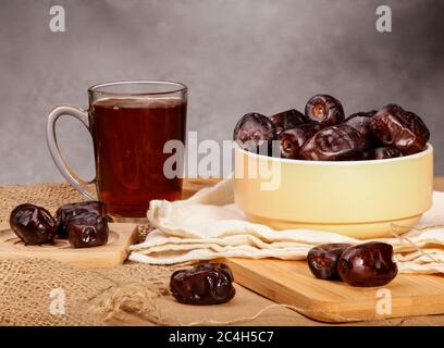 Einige Datteln in einer hellgelben Keramikschale und einige Datteln um sie herum auf einer hölzernen und durchsätderen Oberfläche plus einer Glas Tasse Tee. Ramadan-Datteln und Tee. Stockfoto