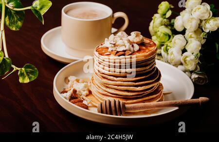 Ein Teller Pfannkuchen Stapel isoliert mit Nüssen, Bananen und Honig, eine Tasse Cappuccino daneben, grün und weiß Blumen um ihn herum auf einem dunklen Tisch. Stockfoto