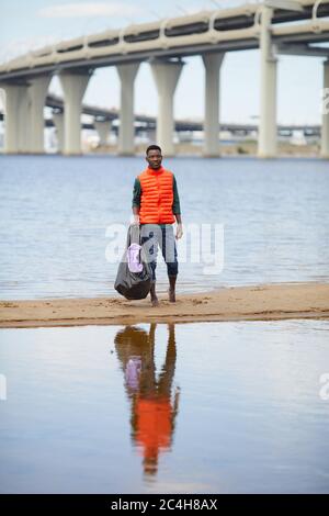 Portrait eines afrikanischen jungen Mannes, der mit Müll am Ufer des Flusses mit einer großen Brücke im Hintergrund steht Stockfoto