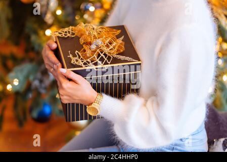 Nahaufnahme der weiblichen Hände mit einem Geschenk (Geschenkbox) in quadratischer Box, weicher Fokus, Weihnachtsbaum auf verschwommenem Hintergrund. Neujahrs-, Geburtstags-Konzept Stockfoto
