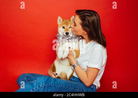 Nette Brünette Frau in weißen T-Shirt und Jeans halten und küssen Shiba Inu Hund in Silber Dekoration auf Ebene roten Hintergrund. Liebe zu den Tieren, pe Stockfoto