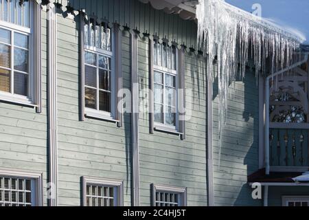 Eis Stalaktit hängen vom Dach mit Holzwand. Gebäude mit großen Eiszapfen bedeckt. Eiszapfen sind sehr gefährlich für das Leben. Schlechte thermische Isolierung Stockfoto