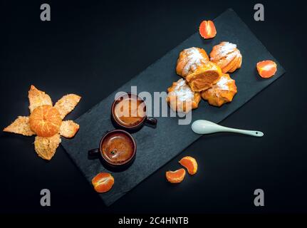 Frischer heißer Kaffee in Keramikbecher, reife Mandarine und süßes Dessert - profiterole oder eclair Kuchen mit Zuckerpulver auf dunkelschwarzem Hintergrund Stockfoto