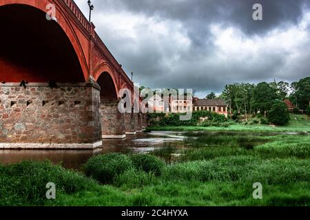 Historische Backsteinbrücke über den Fluss Venta und alte Gebäude in Kuldīga an einem bewölkten Frühlingstag. Stockfoto