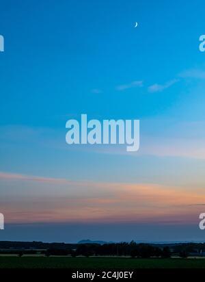 Farbenprächtiger, rosafarbener Himmel in der Abenddämmerung mit Arthur's Seat-Umriss in der Ferne und einem Halbmond am Himmel, East Lothian, Schottland, Großbritannien Stockfoto