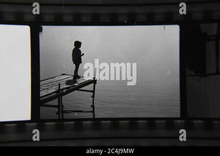 Feine 70er Jahre Vintage-Kontaktdruck Schwarz-Weiß-Fotografie eines Kindes, das am Ende eines Piers steht und auf das ruhige Wasser blickt. Stockfoto