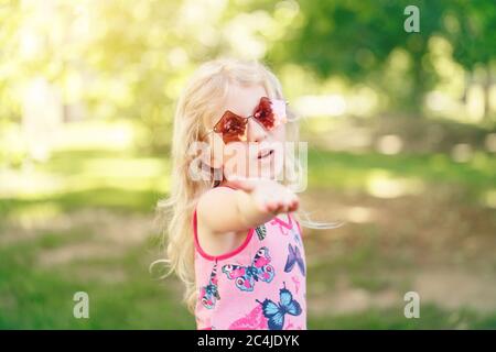 Junges Mädchen posiert in Phantasie rosa fünfeckigen geformten Sonnenbrille im Freien. Cute adorable stilvolle kaukasischen Kind mit langen blonden Haaren bietet Hand Stockfoto