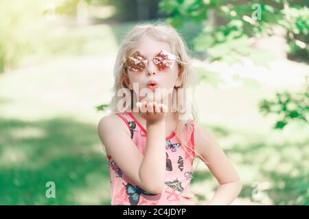 Junges Mädchen posiert in Phantasie rosa fünfeckigen geformten Sonnenbrille im Freien. Cute liebenswert stilvolle kaukasischen Kind mit langen blonden Haaren senden Luftkuss. Co Stockfoto