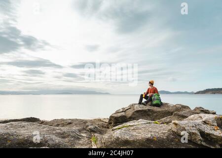 Mann mit Rucksack und auf den Felsen sitzen, während Sie den schönen Morgen am Strand genießen Stockfoto