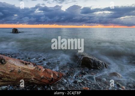 Küstenlinie am Morgen an der Ostsee. Dramatische Wolken bei Sonnenaufgang. Steinige Küste auf der Insel Rügen mit altem Baumstamm und großen Steinen Stockfoto