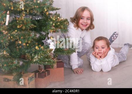 Zwei liebenswerte kleine Schwestern suchen nach Geschenken unter einem Weihnachtsbaum am Heiligabend zu Hause. Schneeflocken-Textur Stockfoto