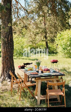 Mehrere Holzstühle um den Tisch mit hausgemachten Speisen und Getränken zum Abendessen im Freien unter Kiefern am sonnigen Sommertag serviert Stockfoto