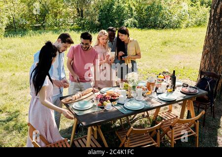 Zwei junge interkulturelle Männer sprechen, während einer von ihnen frisches Baguette auf dem Tisch unter ihren Freundinnen vor dem Abendessen im Freien schneidet Stockfoto