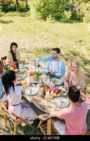 Sechs junge Freunde verschiedener Ethnien, die am Tisch zum Abendessen versammelt sind, unterhalten sich und genießen am Sommertag hausgemachte Speisen Stockfoto