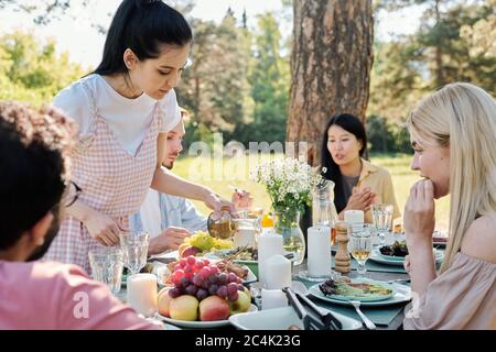 Brunette Mädchen in Casualwear Gießen trinken in Glas, während Biegen von serviert Tisch unter ihren Freunden während Abendessen im Freien Stockfoto