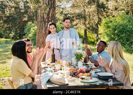 Zwei Paare junger interkultureller Datteln klatschen die Hände, während sie dem amourösen kaukasischen Paar zu ihrem Engagement beim Abendessen gratulieren Stockfoto