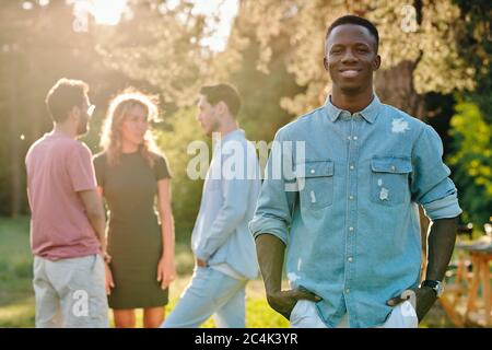 Glücklicher junger Afrikaner im Denim-Hemd, der im Park gegen eine Gruppe seiner interkulturellen Freunde steht, die ihre Ideen darüber diskutieren, wohin sie gehen sollen Stockfoto
