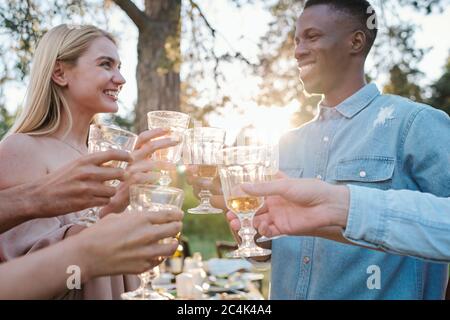 Fröhliche junge interkulturelle Dates, die sich mit toothy Lächeln angucken, während sie mit Gläsern Wein unter ihren Freunden draußen klirren Stockfoto