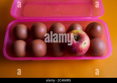 Hühnereier mit Apfel in Plastikbox auf rosa Hintergrund. Konzeptfoto. Stockfoto