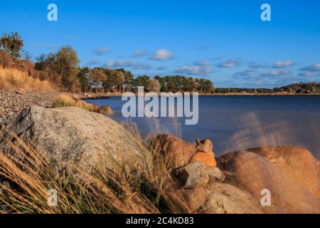 Strandabschnitt auf der Insel Rügen an der Ostsee. Blauer Horizont bei Herbstsonne. Steine und Gräser im Vordergrund. Bäume an der Küste Stockfoto