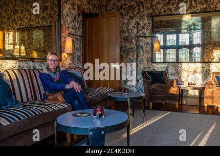 Abendsonne strömt durch Bleiglasfenster in den Damensalon des Hotel Endsleigh, West Devon, England, und fängt handgemalte antike Tapeten mit einem Muster aus Vögeln und Blüten ein. Angela Berg ruht auf einem gestreiften Sofa neben alten Spiegeln, modernen Lampen und ungewöhnlichen Beistelltischen, die zwischen den antiken Sitzgelegenheiten platziert sind. Stockfoto