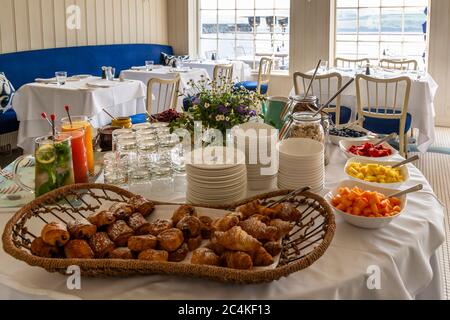 Ein Frühstücksbuffet mit Gebäck, frischem Obst und Getränken ist im Restaurant des Hotel Tresanton in St Mawes, Cornwall, England, aufgebaut. Durch die Bleiglasfenster mit Blick auf den Hafen ist bereits ein Boot sichtbar. Stockfoto