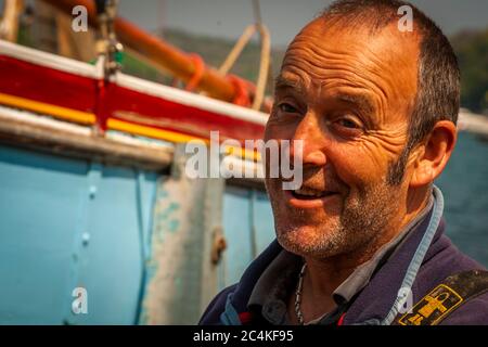 Muschelfischer James Brown lächelt neben seinem traditionellen Arbeitsboot aus Falmouth in der Nähe von St Mawes, Cornwall, England. Er fängt Austern mit der Schute aus dem Fal-Ästuar, wie es die lokalen Vorschriften noch erfordern. Stockfoto