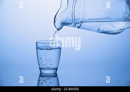 Gießt Wasser aus einem Krug in ein Glas, frisches kaltes Getränk auf blauem Hintergrund Stockfoto