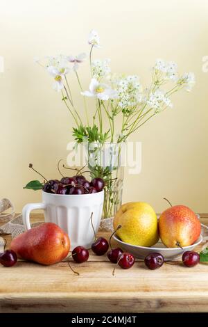 Stillleben mit reifen Birnen, Kirschen und wilden Blumen auf Holztisch. Gemütlicher Hintergrund mit sanfter Morgenbeleuchtung Stockfoto