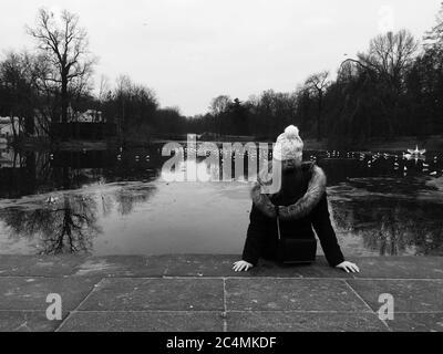 Graustufenaufnahme einer Frau mit einer Mütze auf der sitzt Der Boden ist tagsüber von einem Teich umgeben Stockfoto